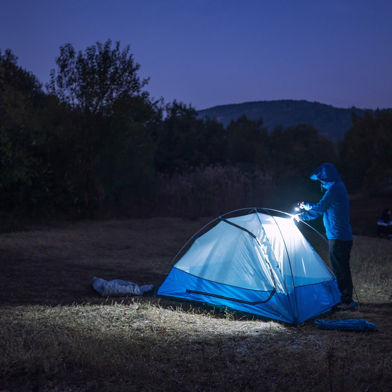 Person setting up a tent in a dark outdoor setting with a flashlight.